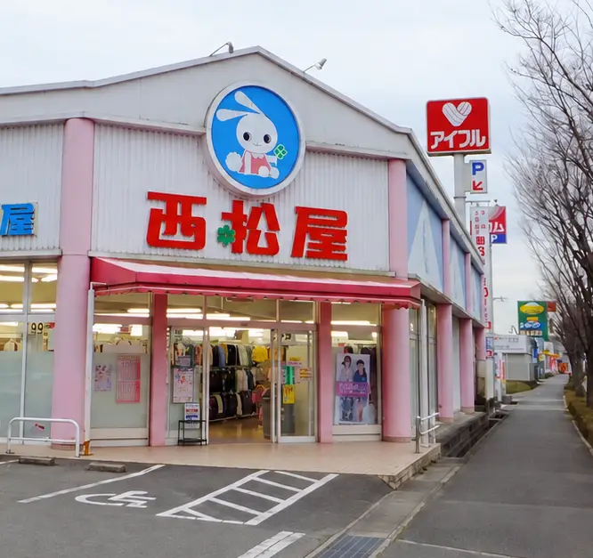 Façade d'un magasin Nishimatsuya avec enseigne au lapin rose, parking en avant et enseignes adjacentes