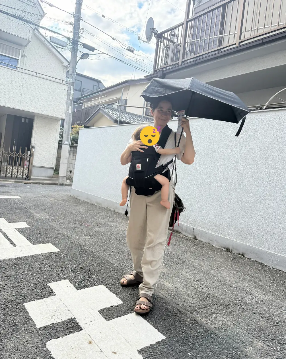 Maman portant bébé endormi dans un porte-bébé Ergobaby, tenant un parasol anti-UV dans une rue résidentielle japonaise