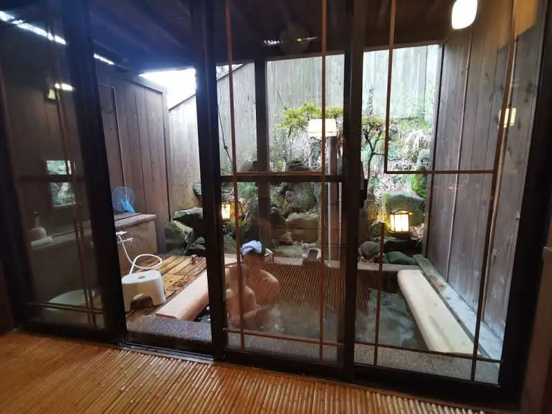 Vue d'ensemble de l'onsen privatif du ryokan Saikatei Jidaiya à Yamagata à travers les portes vitrées, bassin en pierre, coin douche, palissade en bois et jardin de rochers éclairé par des lanternes