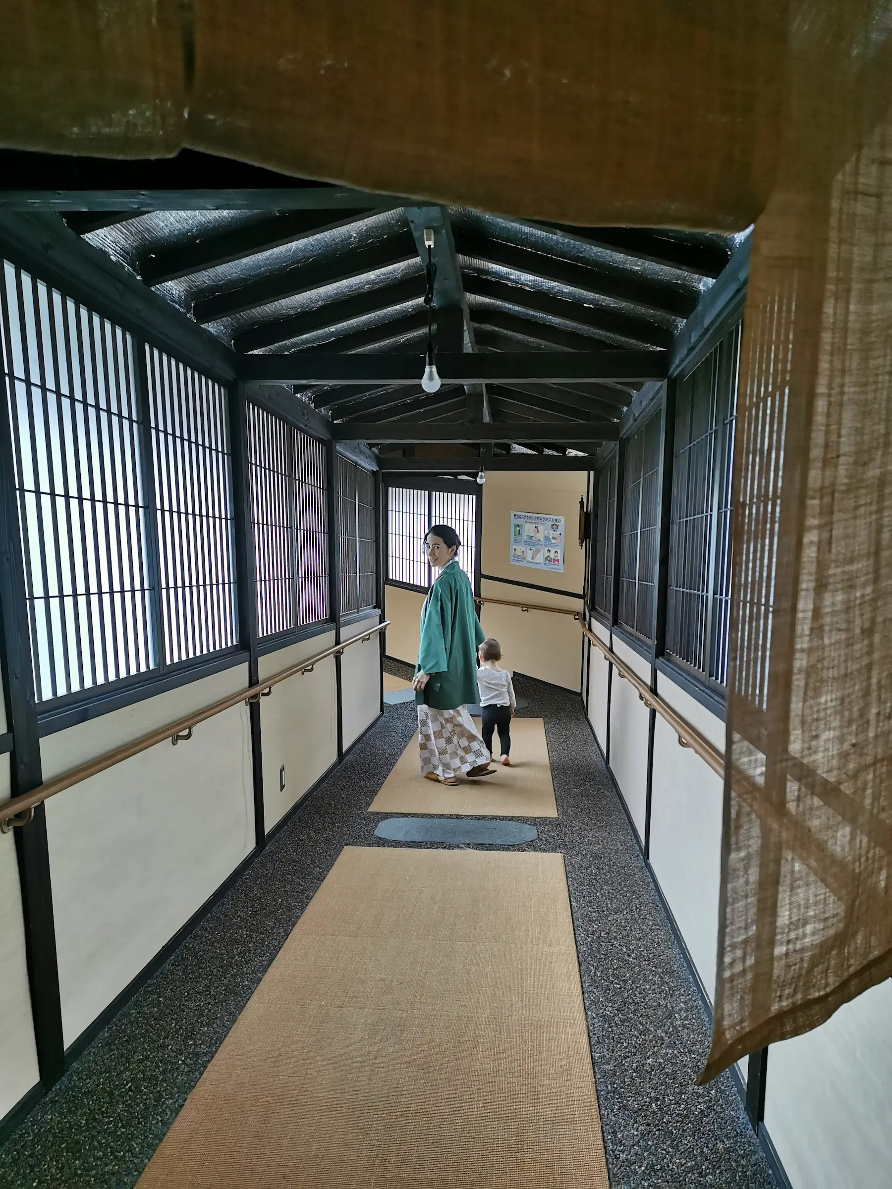 Passage couvert du ryokan Saikatei Jidaiya à Yamagata, charpente en bois sombre et fenêtres à lattes, adulte en yukata et veste hanten marchant avec un jeune enfant